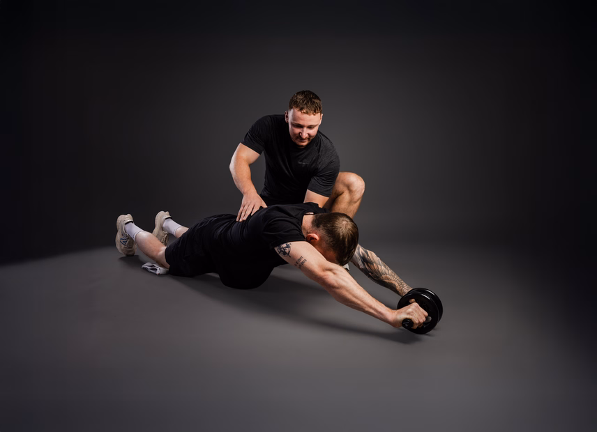 Zwei Männer trainieren in einem Fitnessstudio. Einer liegt auf dem Boden und verwendet ein Hantelrad, während der andere ihn anleitet.
