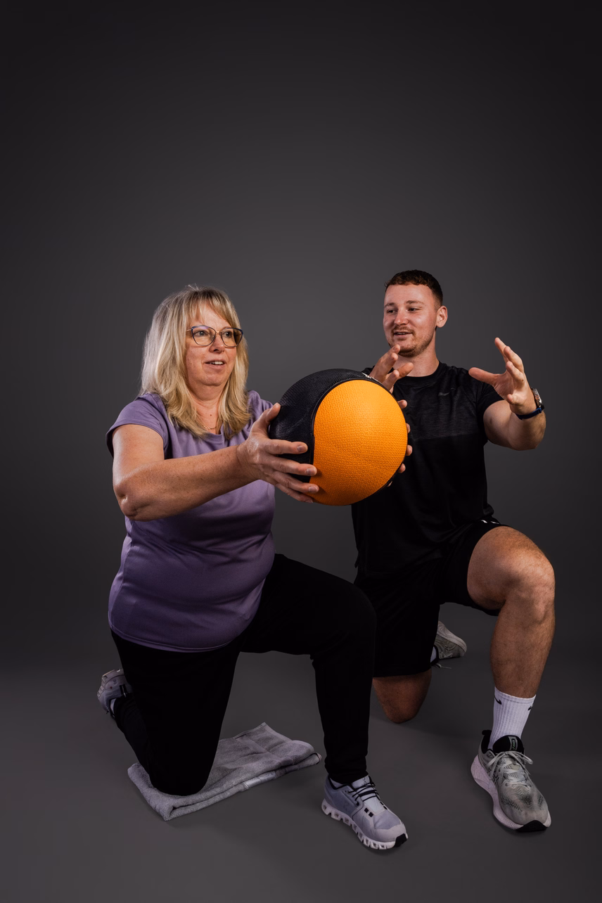 Zwei Personen üben gemeinsam mit einem Medizinball. Eine Frau kniet mit dem Ball, während ein Mann auf einem Knie sitzt und anfeuert. Der Hintergrund ist grau.